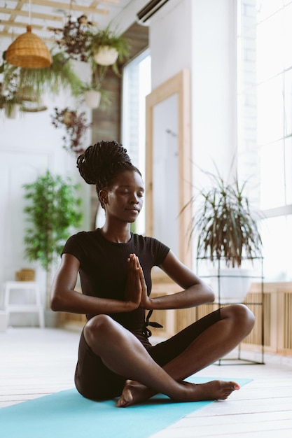 Uma jovem vertical calma e espiritual de pele escura com olhos próximos junta as mãos em namaste Asana sentada em pose de lótus