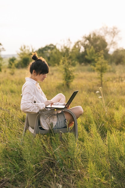 Uma jovem vestindo uma camisa trabalha em um laptop enquanto está sentada em uma cadeira no meio da natureza
