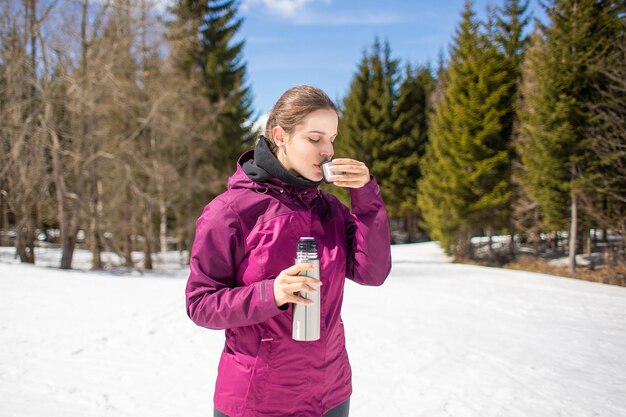 Uma jovem viajante está segurando a garrafa térmica e bebe chá no contexto das montanhas e neve