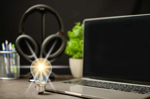 Foto uma lâmpada brilhante e um laptop são uma tela preta em branco na mesa de trabalho conceito de uma startup que impulsiona a economia financeira bancária fones de ouvido sem fio na sala de trabalho fundo preto desfocado