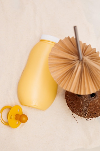 Uma lata amarela de leite em pó está na areia fina com uma chupeta e um coquetel de coco com