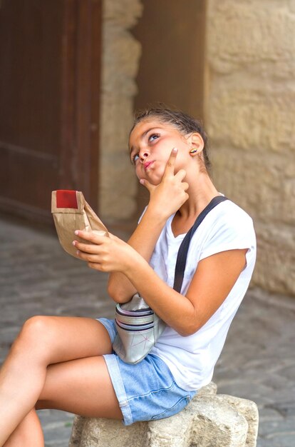 Uma linda adolescente caucasiana de 10 anos está sentada em uma pedra segurando um saquinho de papel com grãos de café nas mãos o que está escrito no rótulo Pensando em comprar um produto