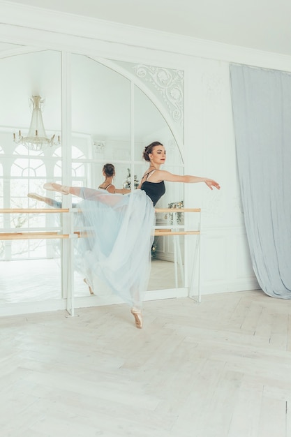 Uma linda bailarina graciosa praticando balé em uma saia tutu azul perto de um grande espelho no corredor de luz branca