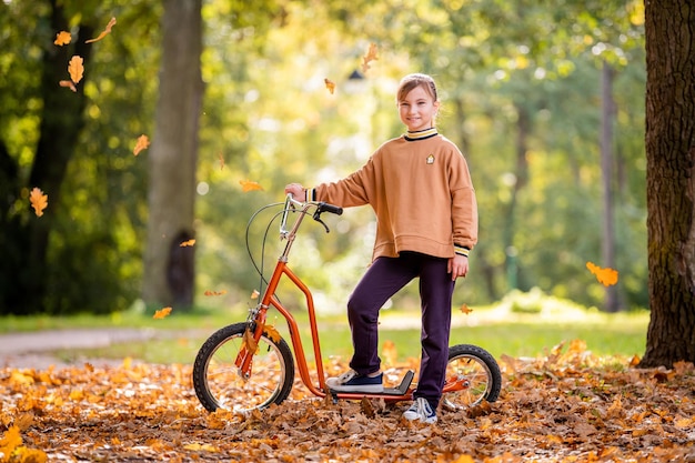 Uma linda colegial anda de scooter em um parque de outono em um dia ensolarado