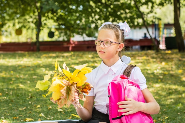 Uma linda colegial com laços brancos está segurando um buquê de folhas de outono e uma mochila em um parque ensolarado de outono. De volta ao conceito de escola. Copie o espaço