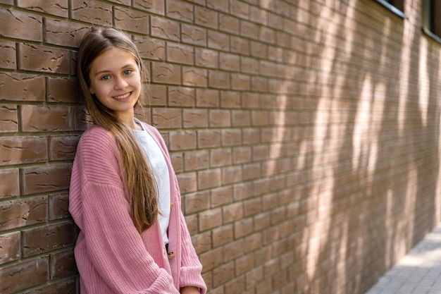 Uma linda colegial está de pé contra uma parede de tijolos no quintal da escola