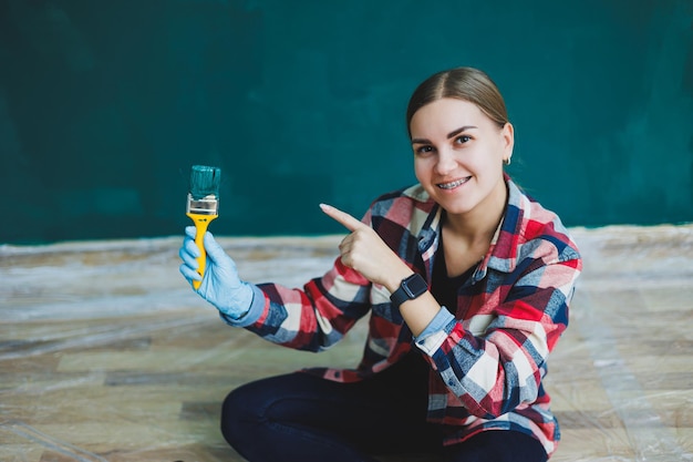 Uma linda designer de mulher faz reparos pinta uma parede cinza com tinta verde com um pincel camisa xadrez cabelo comprido e jeans reparo no apartamento