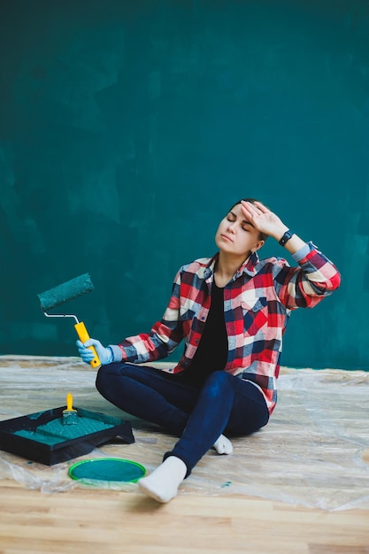 Uma linda designer de mulher faz reparos pinta uma parede cinza com tinta verde com um pincel Camisa xadrez cabelo comprido e jeans Reparo no apartamento