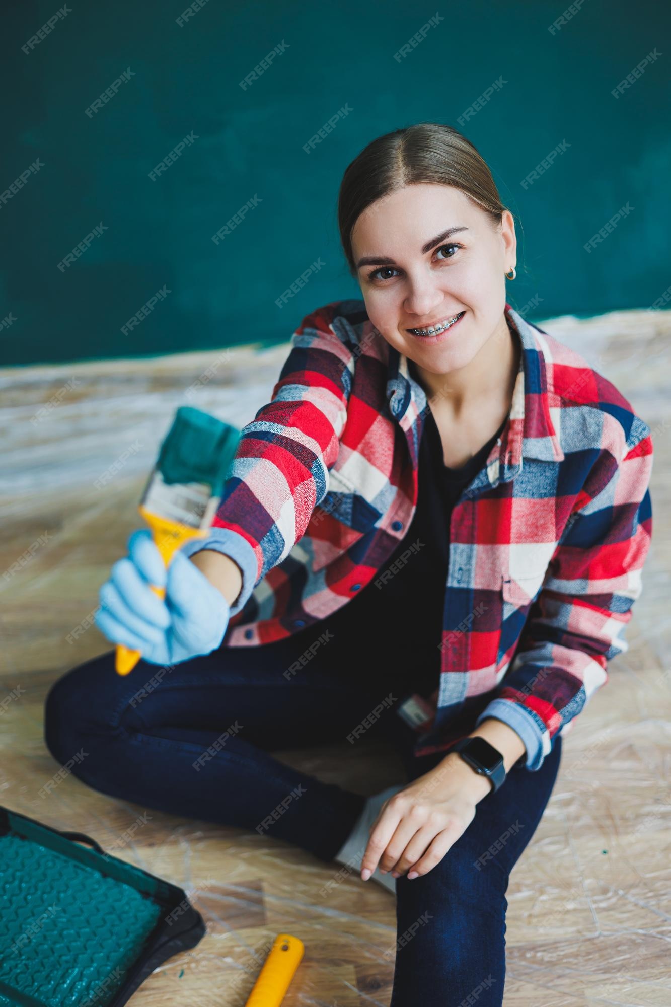 Uma linda designer de mulher faz reparos pinta uma parede cinza com tinta  verde com um pincel camisa xadrez cabelo comprido e jeans reparo no  apartamento