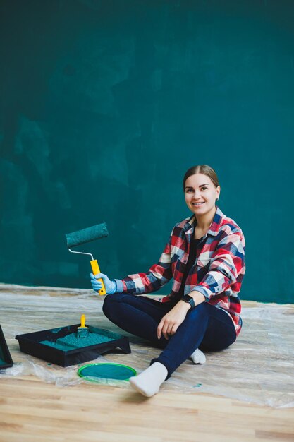 Uma linda designer de mulher faz reparos pinta uma parede cinza com tinta verde com um pincel Camisa xadrez cabelo comprido e jeans Reparo no apartamento