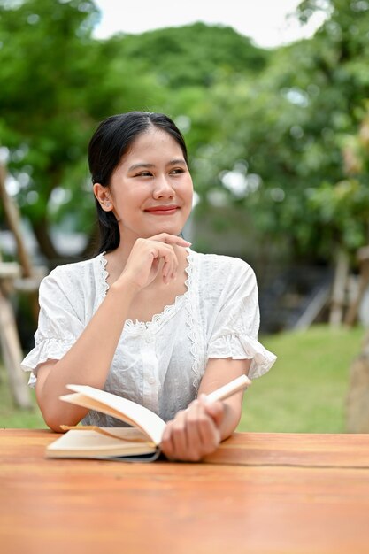 Uma linda e sonhadora mulher asiática está lendo um livro em uma mesa ao ar livre