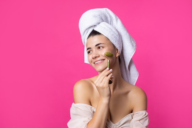 Uma linda garota atraente e alegre com uma toalha na cabeça segura um rolo cosmético perto do rosto contra um fundo rosa