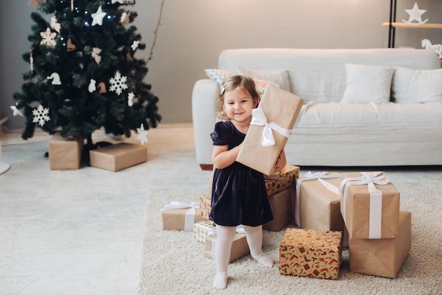 Uma linda garota caucasiana com cabelo loiro curto e ondulado em um lindo vestido preto sentada ao lado de presentes de Natal em uma grande sala iluminada da casa