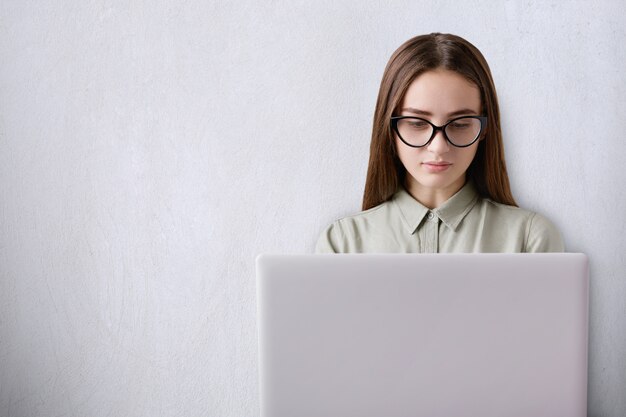 Uma linda garota com cabelos longos, de óculos, sentado na sala e trabalhando com o laptop em branco