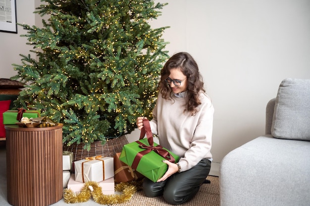 Uma linda garota com óculos está sentada debaixo de uma árvore de Natal com presentes
