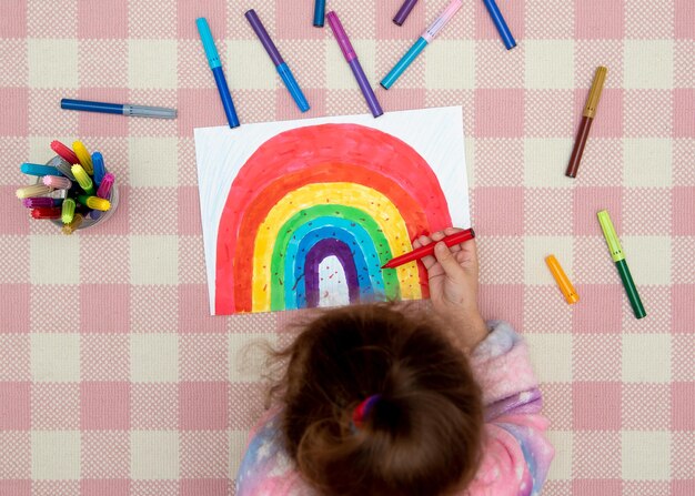 Uma linda garota com roupas de arco-íris desenha um arco-íris com marcadores multicoloridos. Casa. Infância.