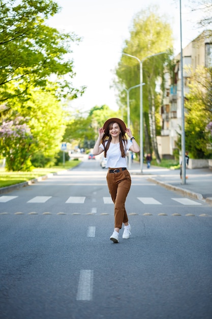 Foto uma linda garota com um chapéu marrom e uma camiseta branca está caminhando na estrada em um dia quente e ensolarado. mulher jovem e bonita com um chapéu com um sorriso no rosto