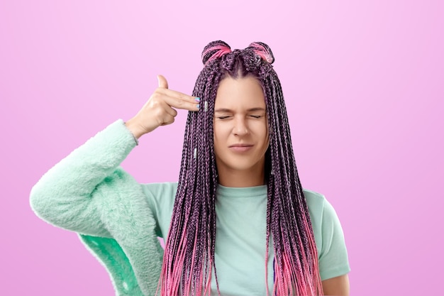 Foto uma linda garota com um penteado de tranças rosa em roupas turquesa dá um tiro na cabeça com os dedos. o conceito de fadiga, obsessão, negatividade.