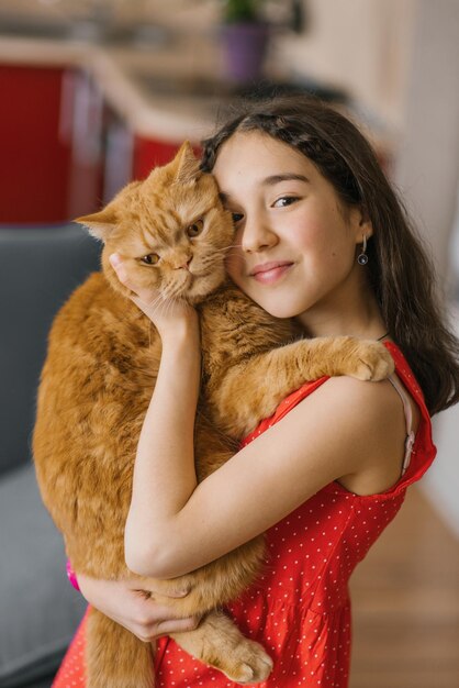 Foto uma linda garota com um vestido vermelho segura seu amado gato britânico vermelho em seus braços