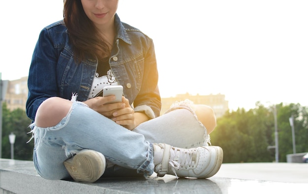 Uma linda garota com uma jaqueta jeans sentada nos degraus de uma rua da cidade usa seu smartphone