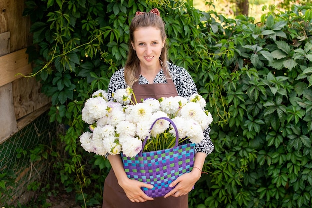 Uma linda garota de avental com uma cesta de flores de dálias brancas está de pé perto de um vinhedo