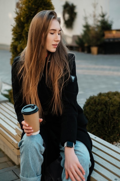 Uma linda garota de cabelo comprido com café sentado em um banco e olhando para longe Retrato feminino de rua casual