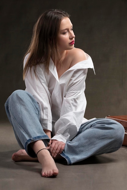 Uma linda garota de camisa branca e jeans senta-se em um fundo cinza