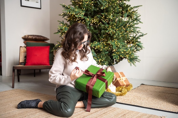 Uma linda garota de óculos está sentada no tapete perto da árvore de Natal com presentes