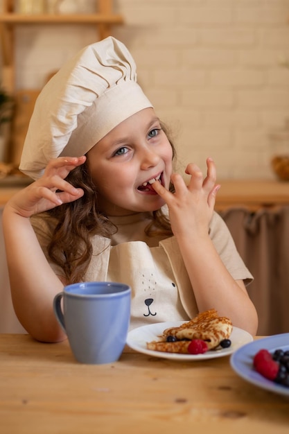 uma linda garota de seis anos com chapéu e avental de chef está sentada em uma mesa de madeira comendo panquecas