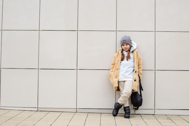Uma linda garota de seis anos com um casaco de pele falso bege está de pé contra uma parede na cidade
