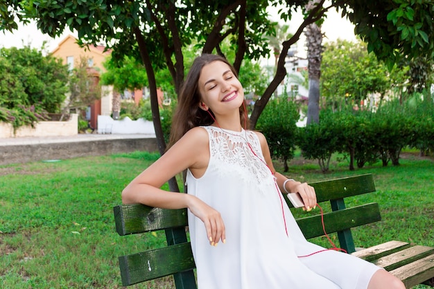 Uma linda garota de vestido branco está ouvindo sua música favorita no banco do parque. Ela gosta e está se divertindo.