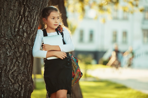 Uma linda garota em idade escolar está de pé ao lado de uma árvore e segurando um tablet. Colegial vestida com um vestido de verão