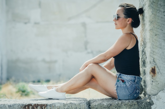 Uma linda garota em shorts jeans e uma regata preta sentada encostada na parede