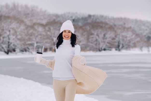 Uma linda garota em um cardigã bege e um chapéu branco com um copo de chá desfruta de um aterro nevado à beira do lago