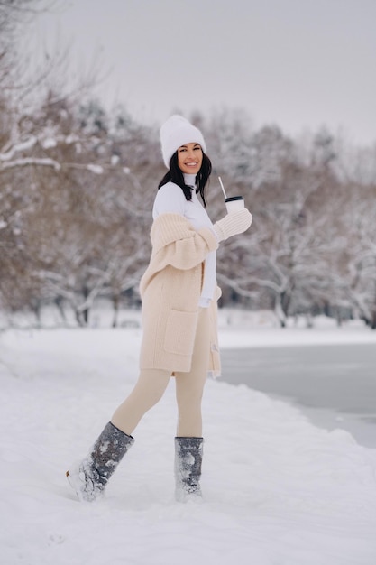 Uma linda garota em um cardigã bege e um chapéu branco com um copo de chá desfruta de um aterro nevado à beira do lago