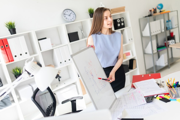 Uma linda garota em um escritório fica perto de uma mesa e explica em um quadro negro magnético um cascalho.