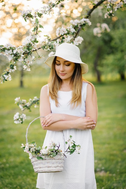 Uma linda garota em um jardim florescente segura uma cesta com flores de maçã nas mãos