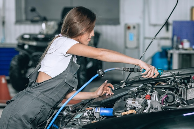Uma linda garota em um macacão preto e uma camiseta branca está sorrindo, verificando o nível de óleo em um carro preto na garagem.