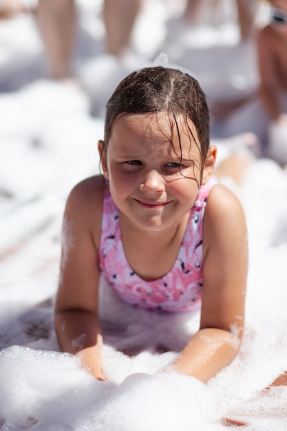 Uma linda garota em um maiô rosa encontra-se sob a espuma em uma festa de espuma na praia à beira-mar no verão ...