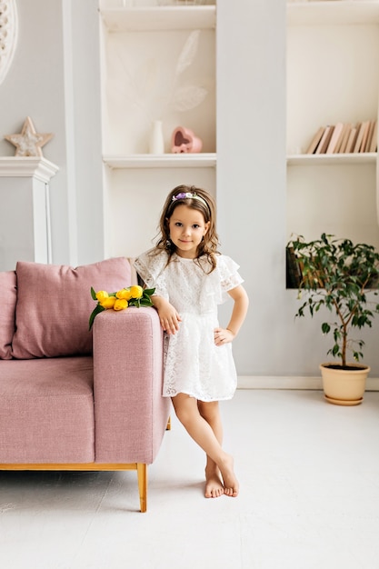 Uma linda garota em um vestido branco com um buquê de tulipas está ao lado dela no sofá da sala.