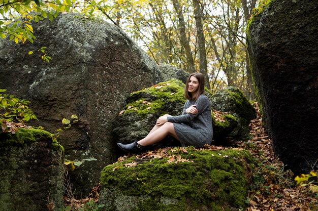 Uma linda garota em um vestido cinza senta-se no outono em uma pedra coberta de musgo na floresta e sorri agradavelmente.