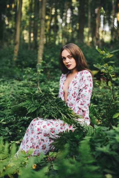 Uma linda garota em um vestido floral está sentado com um buquê de samambaia na floresta.