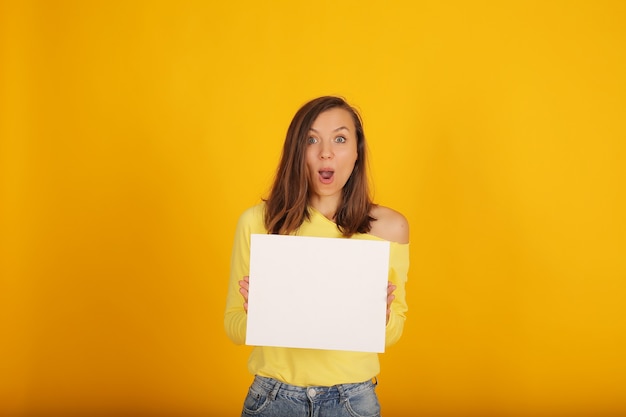 uma linda garota em uma jaqueta amarela e jeans segura um pedaço de papel branco sobre um fundo amarelo