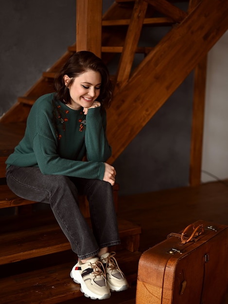 Uma linda garota está sentada ao lado de uma mala em uma escada de madeira e olha pela janela