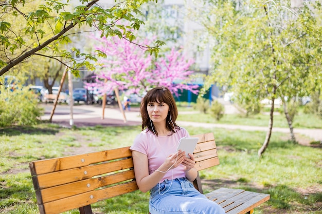 Uma linda garota está sentado em um banco com um tablet, fones de ouvido e parece longe. uma jovem garota em um top rosa e jeans está sentado em um banco na rua.