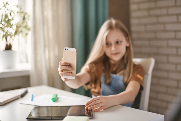 Uma linda garota faz uma pausa na lição de casa e tira uma selfie em seu telefone móvel, sentado à mesa.