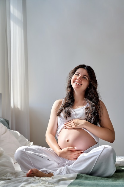 Uma linda garota grávida segura a barriga com as mãos e sorri.