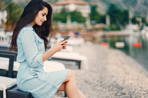 Uma linda garota jovem em um vestido azul, sentado perto da água e usar o telefone