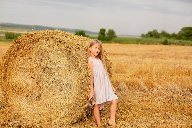 uma linda garota loira com cabelos longos em um vestido de linho fica por um palheiro em um campo ceifado