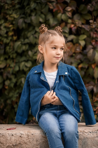 Uma linda garota loira elegante com uma jaqueta azul se senta em um bloco de concreto. Menina de 7 anos com um sorriso em um fundo de folhas